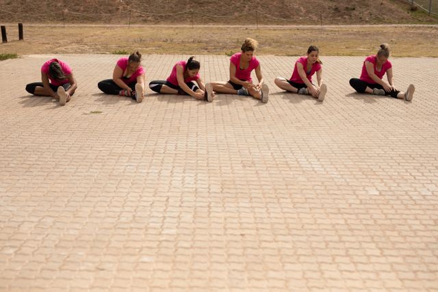 Women in pink shirts stretching legs during outdoor boot camp training session. Ideal for promoting group fitness activities, outdoor exercise programs, healthy lifestyle campaigns, and team-building events.