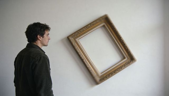 Standing man observing tilted gilded frame on blank wall for minimalist contemplation