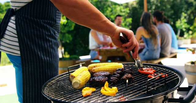 Man grilling vegetables and meats on a barbecue grill during a casual summer gathering. Corn, peppers, and other delicacies are being prepared with tongs while friends are socializing in the background. Ideal for summer events, outdoor cooking articles, and social gathering promotions.