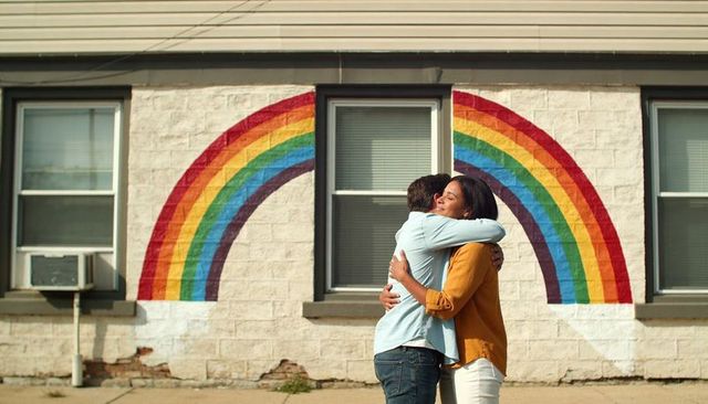 Sunlit couple embracing under rainbow mural on urban sidewalk