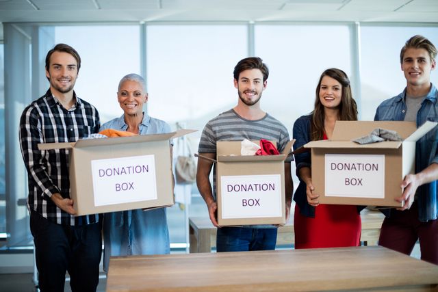 Group of colleagues holding donation boxes filled with clothes in an office setting. Ideal for use in articles or campaigns related to corporate social responsibility, charity events, community service, and team building activities.