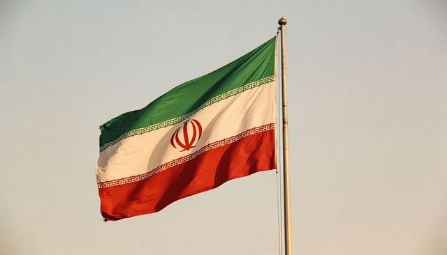 Iranian flag waving at dusk on tall pole with gold finial, emblem and tricolor fabric