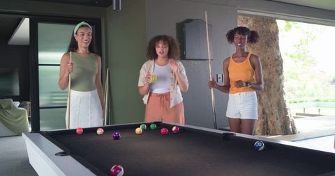 African american friends playing pool on black felt table in sunlit modern game room