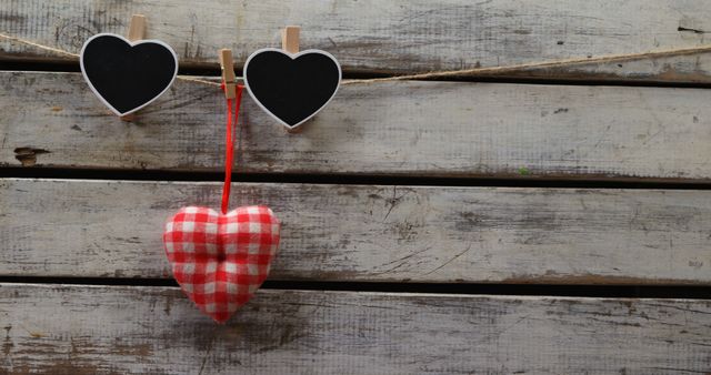 A red and white checkered heart ornament hangs between two heart-shaped blackboards clipped to a string, with copy space. Evoking a sense of love and celebration, the rustic wooden background adds a charming, vintage feel to the composition.