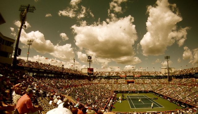 Large audience fills the seatings in a tennis stadium during an event on a sunny day with scattered clouds in the sky. Suitable for promoting sports events, tournament advertisements, and tennis-related articles.