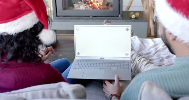 Couple wearing Santa hats using a laptop by a fireplace, emphasizing a sense of cozy winter holiday season. Perfect for prints or digital ads featuring themes of holiday advertising, e-commerce, or social media during Christmas. Ideal for promotions highlighting tech products or Christmas sales in a homey, warm setting.