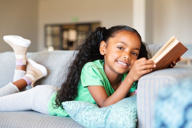 Portrait of smiling african american elementary girl holding book while lying on sofa in school. unaltered, childhood, education, reading, relaxation and school concept.