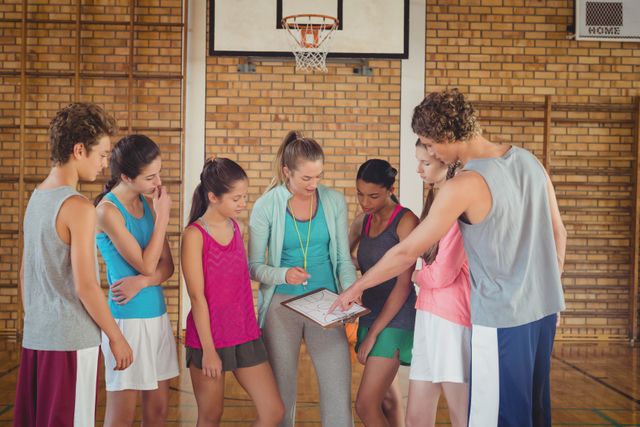 Female coach mentoring high school basketball team in gym. Students gathered around coach, listening and discussing strategy. Ideal for use in educational materials, sports training guides, teamwork and leadership articles, and youth development programs.