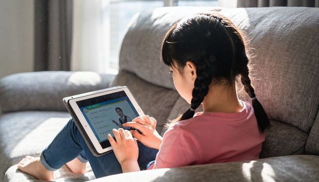 Young girl studying on tablet on sunlit sofa at home watching online lesson edtech learning