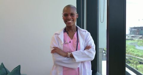 African woman doctor smiling with arms crossed in clinic by window wearing scrubs and lab coat