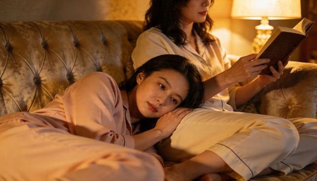 Women in pajamas relaxing on velvet tufted sofa reading together under warm lamp light
