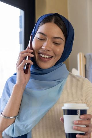 Young woman in blue hijab talking on phone holding takeaway coffee by window casual morning