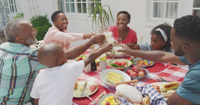 Happy african american family sitting at table in garden, eating dinner and making a toast. Lifestyle, domestic life, family, and togetherness.