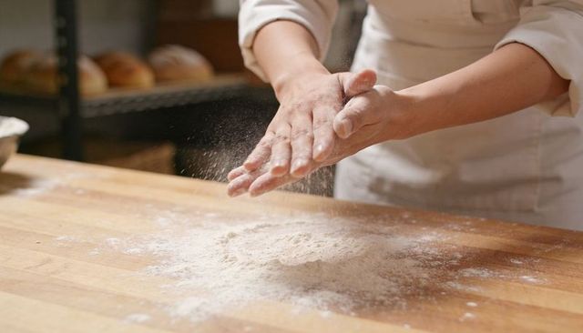 Baker clapping flour from hands over wooden counter, flour dust cloud in bakery