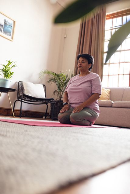Low angle view of biracial mature woman with eyes closed kneeling and meditating on mat at home. Unaltered, exercise, yoga, retirement, zen, healthy and active lifestyle concept.