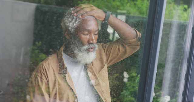 Elderly man with gray beard thoughtfully gazing through a window, showing reflection of outdoor calm scene. Dressed in casual attire with wristwatch, conveying a contemplative mood. Perfect for themes of mindfulness, introspection, mental health awareness, solitude, and peaceful living.