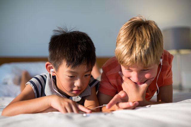 Two children are lying on a bed at home, sharing earphones and using a mobile phone. This image can be used to depict themes of technology, childhood bonding, leisure activities, and family time. It is suitable for use in advertisements, blogs, and articles related to family life, technology use among children, and home activities.