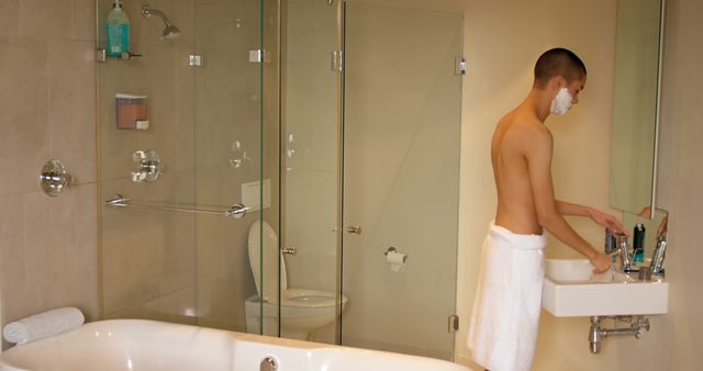 Shirtless man with shaving cream on his face stands at the sink in a modern bathroom, ready to shave. Useful for topics related to male grooming, personal hygiene, self-care routines, modern home interiors, and daily lifestyle habits.