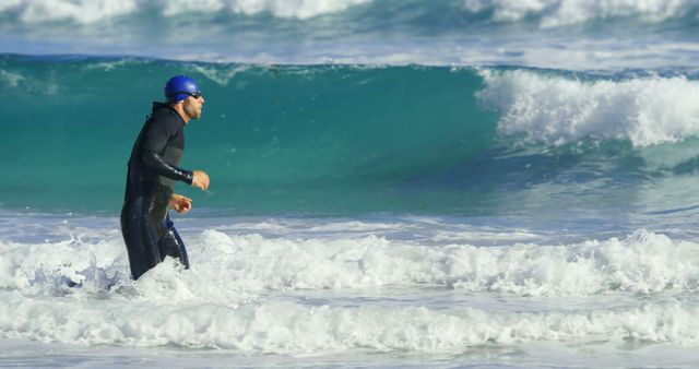 A middle-aged Caucasian man in a wetsuit and swim cap is running through the surf on a beach. His focused expression and athletic gear suggest he might be training for a triathlon or engaging in open water swimming exercises.