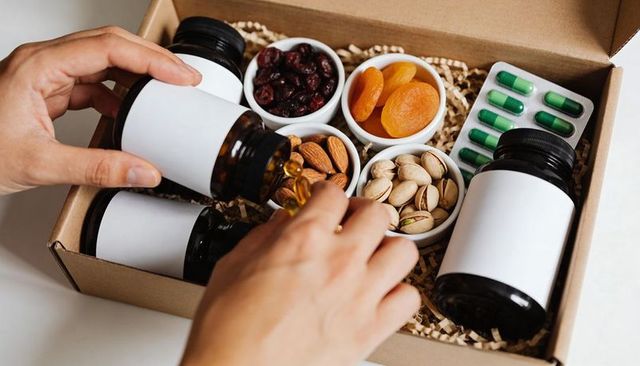 Pouring softgels into wellness gift box with supplement bottles, nuts, dried fruit flatlay
