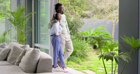 Indian and african american couple standing at sliding door enjoying lush garden view