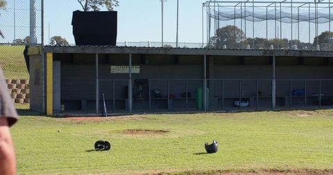 Male standing on outfield edge observing dugout with helmets scattered on green grass