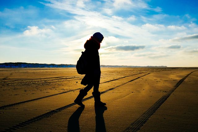 Silhouette of individual walking on beach during sunset with tracks in sand and backpack. Suitable for travel blogs, adventure websites, or inspirational posts. Emphasizes themes of exploration, solitude, and natural beauty.