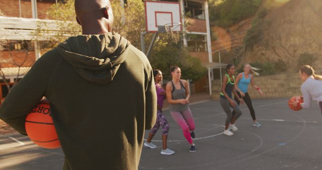 A coach holding a basketball is watching a group of women engaged in an outdoor basketball practice. The participants are energetically active, working on their basketball skills and fostering team spirit. Useful for promoting teamwork, active lifestyles, sports coaching, and women's athletic events.