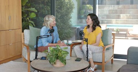 Two women recording conversational podcast at home living room with microphones and laptop