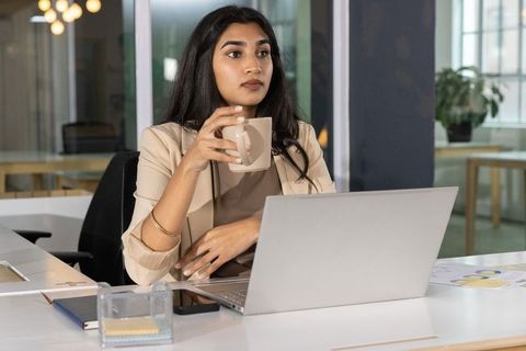 Indian businesswoman holding coffee while working on laptop in modern open-plan office