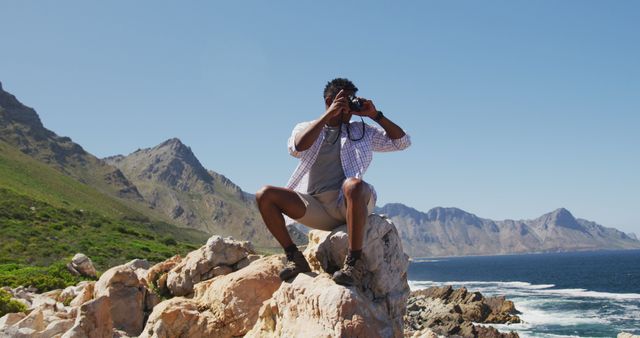 African american man hiking sitting on rock taking photos by the coast. fitness training and healthy outdoor lifestyle.