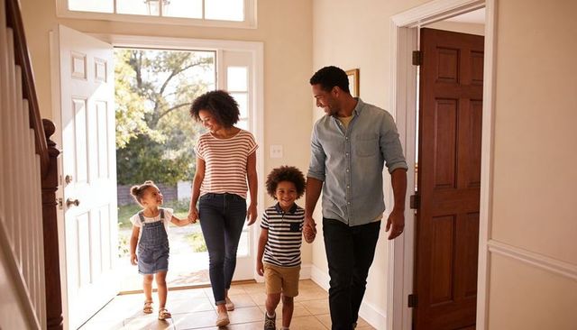 Family arriving home smiling in sunlit foyer, parents holding hands with children, casual outfits
