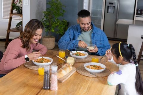 Smiling family enjoying pasta dinner around wooden table in modern cozy open-plan kitchen