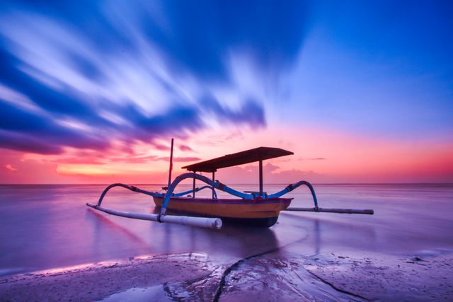 Traditional boat anchored in calm ocean during vibrant sunset. Creative long exposure captures dynamic clouds across colorful sky, reflecting on smooth water. Ideal for travel marketing, inspirational posters, coastal living decor, and serene lifestyle promotions.