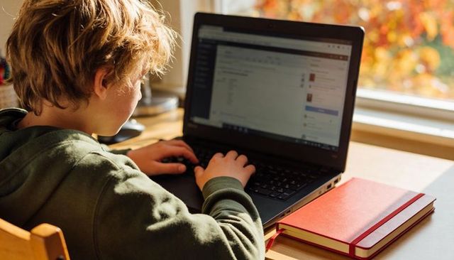 Young student typing on laptop at sunlit desk with red notebooks and autumn window