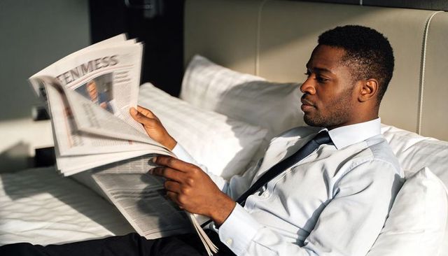 Business traveler reading broadsheet newspaper on hotel bed, relaxing in shirt and tie