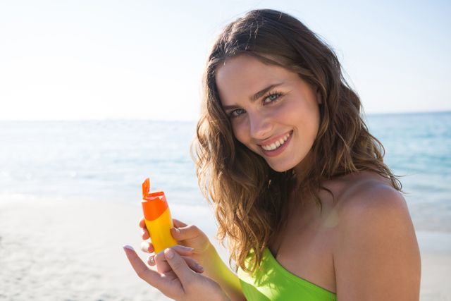 Young woman enjoying a sunny day at the beach, holding sunscreen and smiling. Ideal for use in advertisements for skincare products, summer vacation promotions, health and wellness campaigns, and travel brochures. Highlights the importance of sun protection and enjoying outdoor activities.