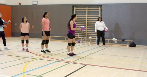Diverse female volleyball team practicing passing drill in indoor gym with coach guiding