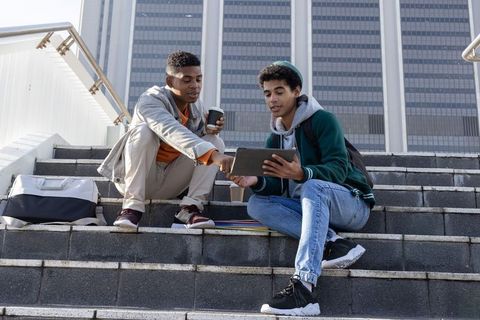 Diverse teenage friends sharing tablet and coffee while studying on city steps