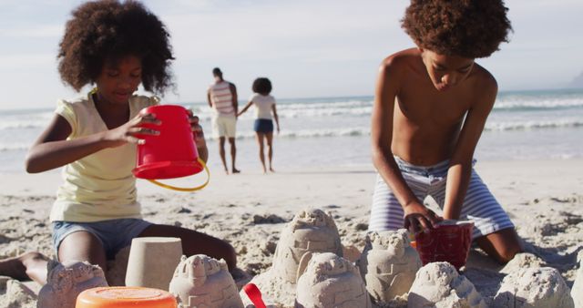 Children enjoying a sunny day at the beach are building sandcastles with red buckets and fine sand. Family members can be observed in the background, making it a perfect scene that encapsulates summer vacation fun. This can be ideal for promotions related to family vacations, beach resorts, summer activities, and outdoor family fun.