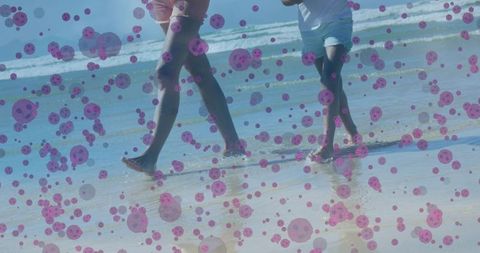 Mother and daughter walking barefoot on beach splashing water with purple bokeh overlay