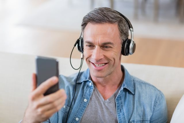 Middle-aged man wearing headphones, smiling while looking at mobile phone in living room. Ideal for use in advertisements for music streaming services, mobile apps, technology products, or lifestyle blogs focusing on relaxation and home entertainment.