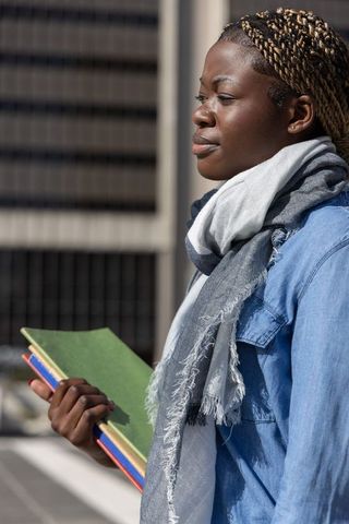 Sunlit african american student holding notebooks in denim jacket and scarf, city portrait