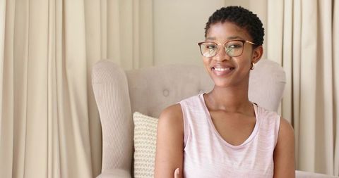 Smiling woman relaxing in wingback chair wearing glasses and pink top, minimalist interior