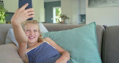 Preteen girl reclining on couch taking selfie with smartphone in bright living room