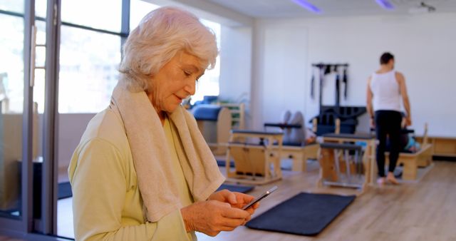 Elderly woman standing in a gym checking her smartphone, showing modern technology use among seniors. Active person in the background by fitness equipment. Useful for promoting health, fitness, workout routines, active lifestyles for seniors, technology adoption among older adults.