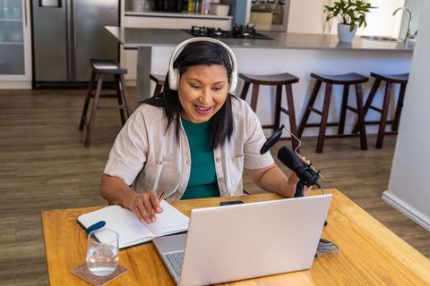 Mature woman podcasting from home kitchen with headset, laptop, notebook and microphone
