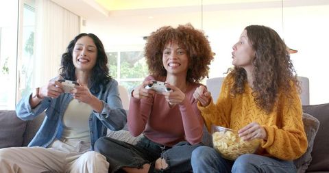 Female friends playing multiplayer console game on couch holding controllers sharing chips