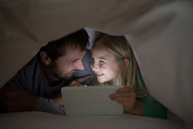 Father and daughter lying under blanket and using digital tablet at home