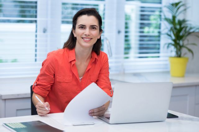 This image shows a woman smiling while writing on paper at her home office. She is seated at a desk with a laptop and other office supplies. The setting is bright with natural light coming through the window, and a plant adds a touch of greenery. This image can be used for promoting remote work, productivity, home office setups, and professional business environments.